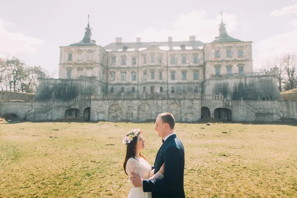 Attractive newlywed couple embracing at green sunny lawn near beautiful ruined baroque palace. Loving groom holding charming bride in his hands