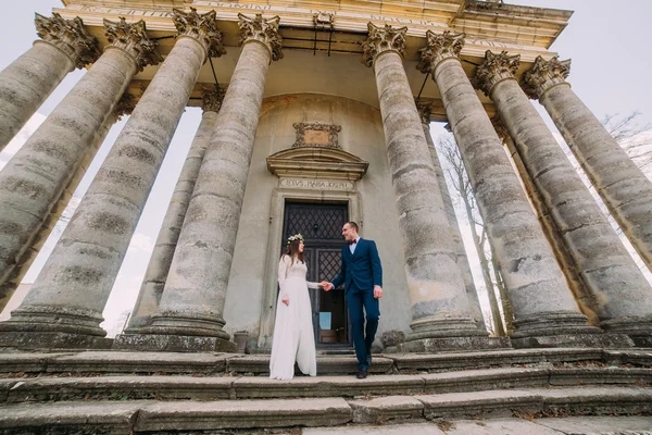 Beautiful newlywed couple go down by stairs of the old victorian church with columns