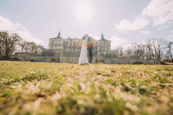 Attractive newlywed couple embracing at green sunny lawn near beautiful ruined baroque palace. Loving groom and charming bride touching by foreheads