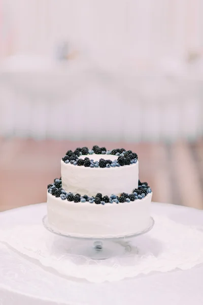 Candid cream cake decorated with bilberries and blackberries isolated close-up on table covered by white tablecloth at blurred bright background of restaurant interior