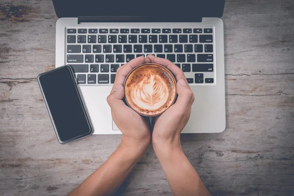 Business man hands holding cup of coffee with latte art on wooden laptop computer and smart phone background, Top view, Vintage filter