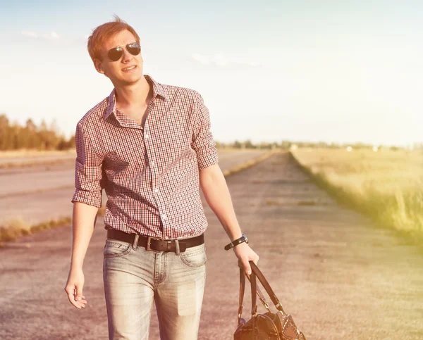 Stylish man in the jeans and sunglasses with the bag walking on the road outdoor