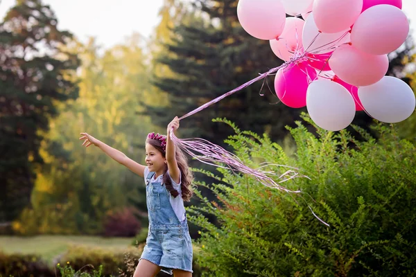Laughing beautiful girl in denim overalls  run on a green lawn with a big bunch of pink  and white balloons.