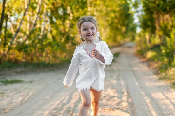 A smiling little girl in a white traditional ornamented chemise running in a scenic early autumn landscape along a country road