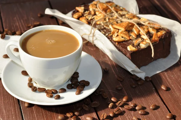 A photo of an an appetizing chocolate cake decorated with small pieces of biscuits and placed wrapped in a rustic paper and a cup of milk coffee