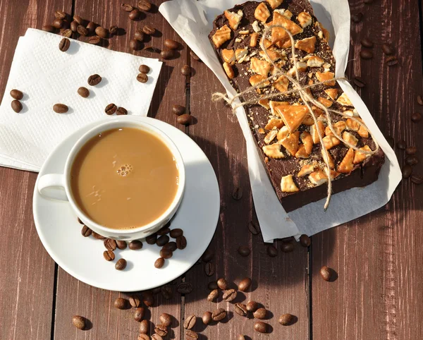 A photo of an an appetizing chocolate cake decorated with small pieces of biscuits and placed wrapped in a rustic paper and a cup of milk coffee