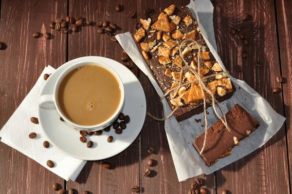 A photo of an an appetizing chocolate cake decorated with small pieces of biscuits and placed wrapped in a rustic paper and a cup of milk coffee