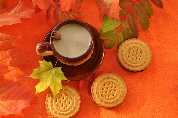 A composition representing and autumn concept -  a cup of milk coffee, a cinnamon stick, biscuits and colorful autumn leaves on a bright orange soft fabric surface
