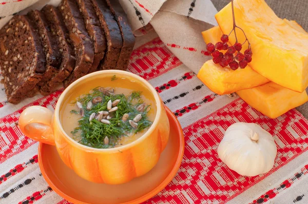 An orange cup of pumpkin soup, several slices of rye bread, pieces of fresh pumpkin and garlic on a linen surface decorated with traditional slovenic embroidery