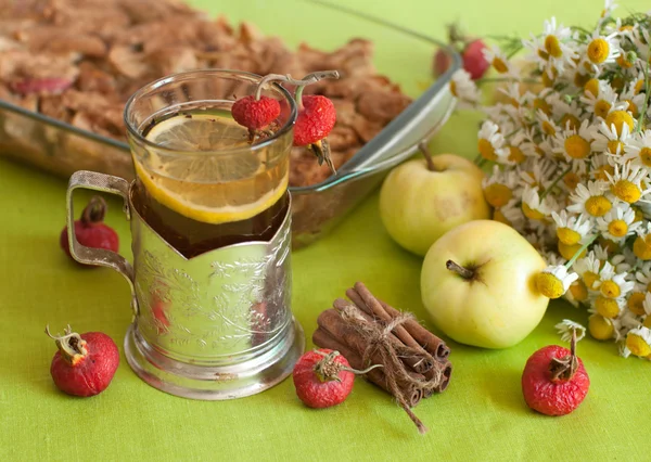 A cup of strong black tea with a lemon slice, an apple pie, a bouquet of chamomiles, cinnamon sticks, ripe apples and mature hips on a light green linen surface