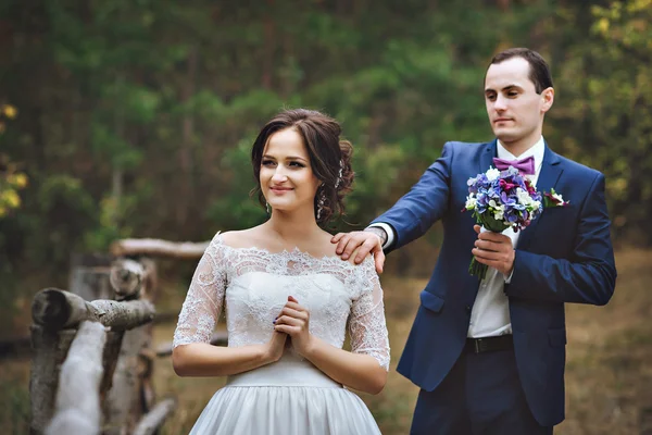 The groom stands behind the bride looks at her, touches the shoulder of his wife. future wife in a lace dress in the foreground, in the woods.