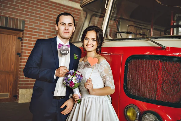 Happy beautiful couple of lovers on a background of a red bus with purple lilac bouquet in her hands. In the hands of decorative props on sticks.