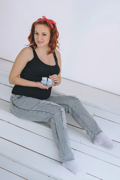 Happy pregnant woman holding booties, with a red bandage on his head. On a light background. Pregnancy red-haired young woman in a black t-shirt.