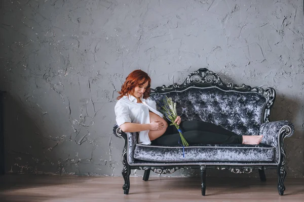 Pregnant happy young woman lying on sofa in the Baroque style. In her hands the flowers. Redhead pregnant woman in white shirt with bare tummy. Looking at the belly. There is a place for the text.