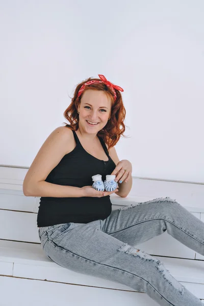 Happy pregnant woman holding booties, with a red bandage on his head. On a light background. Pregnancy red-haired young woman in a black t-shirt.