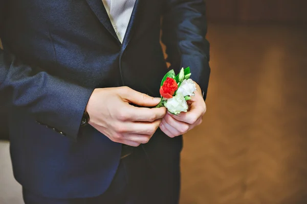 Mens blue suit, red tie, white shirt. The groom adjusts his boutonniere of roses.