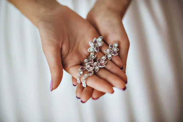 Beautiful bride in white wedding dress is holding earrings in her hands. Beauty model girl in bridal gown for marriage. Female portrait with jewelry. Close-up womans arms. Cute lady indoors