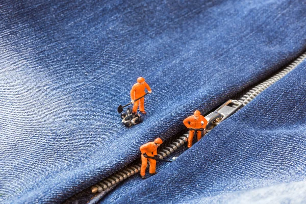 The concept for the fashion industry. Miniature workers sew on buttons big needle to jeans, close up.