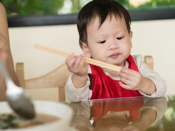 Asian toddler learn to eat meal herself holding chopsticks.