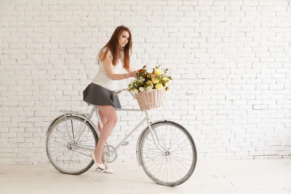 Lovely girl with a bike and basket of flowers on a background of a white brick wall
