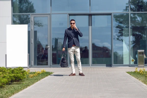 Young official man expert calling via cell telephone while standing outdoors near modern glass building,  male employer talking on mobile phone