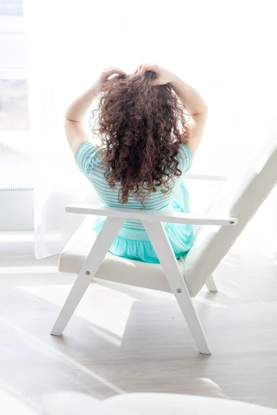 Portrait of a girl with curly hair relaxing in a chair in the morning before a big bright window. Beautiful tender woman in a turquoise dress sitting with his back to the camera.