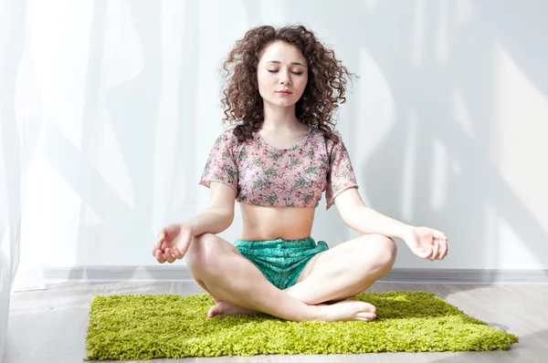 Young girl with curly hair sitting on the floor, on a green rug, do stretching exercises. Doing exercises in the morning in the bright sun room. Meditation. Practice yoga at home. Enjoy life.