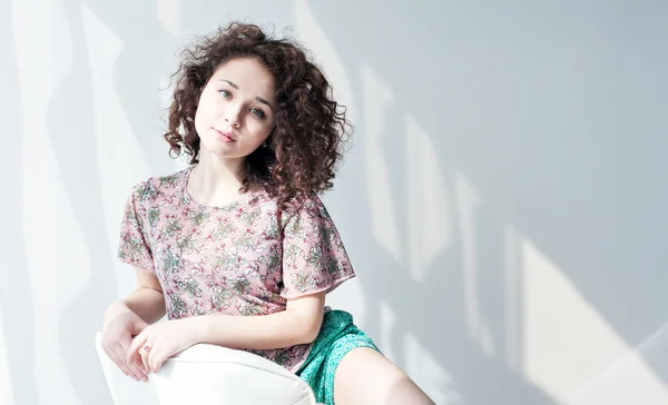 Portrait of a young beautiful girl with curly hair sitting in a white chair in a bright sunny room. Brown wavy hair. Enjoy life.