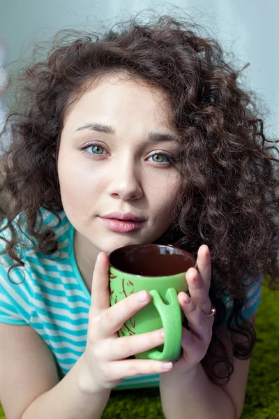 Portrait of a beautiful young girl with brown curly hair. Drink your morning coffee. Girl with a cup of tea in his hands looking at the camera. Big green eyes and green cup in his hands.