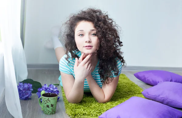 Portrait of a beautiful young girl with brown curly hair. Drink your morning coffee. Girl lying on the floor on a mat for meditation and looking at camera. Big green eyes. Pillows purple.