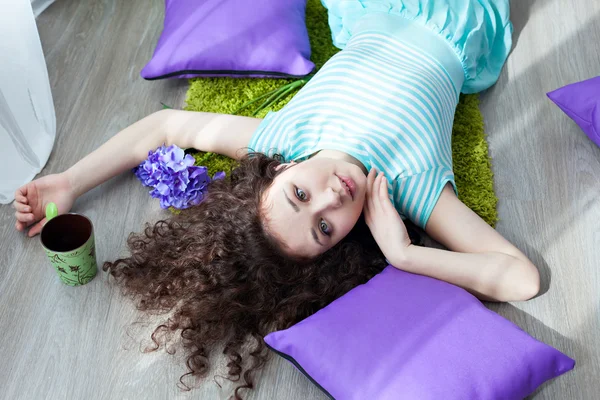 Portrait of a beautiful young girl with brown curly hair. Drink your morning coffee. Big green eyes. Pillows purple. Girl lying on the floor on a mat for meditation and looking at camera.