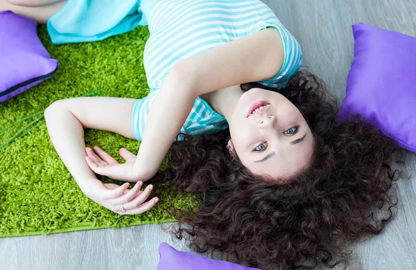 Portrait of a beautiful young girl with brown curly hair. Girl lying on the floor on a mat for meditation and looking at camera. Big green eyes. Pillows purple. Drink your morning coffee.