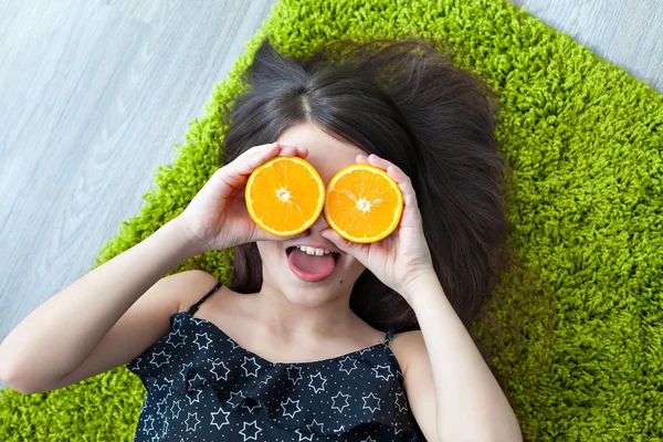 Little girl lying on the floor and holding a slice of orange as the orange glasses. View from above. Summer mood.