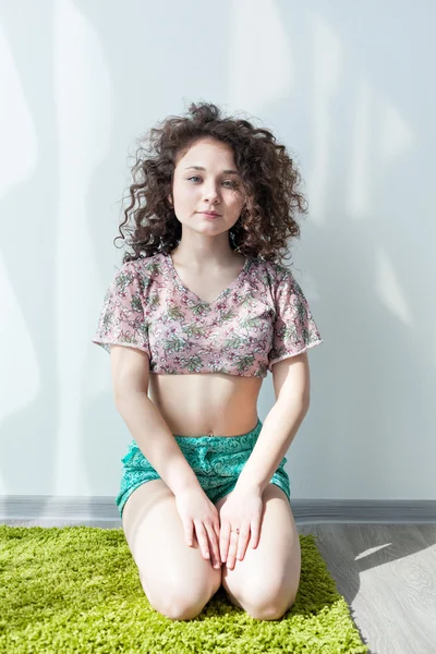 Young girl with curly hair sitting on the floor, on a green rug, do stretching exercises. Practice yoga at home. Enjoy life. Doing exercises in the morning in the bright sun room. Meditation.