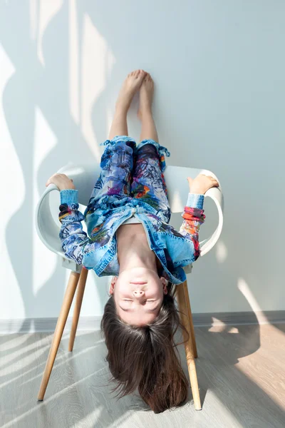 Portrait of a little girl of fashion baby on a white chair. Stylish little girl with long hair is sitting upside down, hanging his head down. Photo model child cool fashionable. Carefree fun