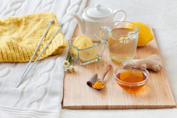Herbal tea in glass cup with flowers of chamomile, turmeric and honey on a wooden board. Treatment with a cold drink. Treatment of folk remedies in bed. Knitting hobby.