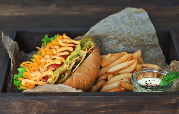 Big tasty hot dog with sausage and vegetables with sauce and French fries on the parchment on the wooden background. hot dogs to gourmet.