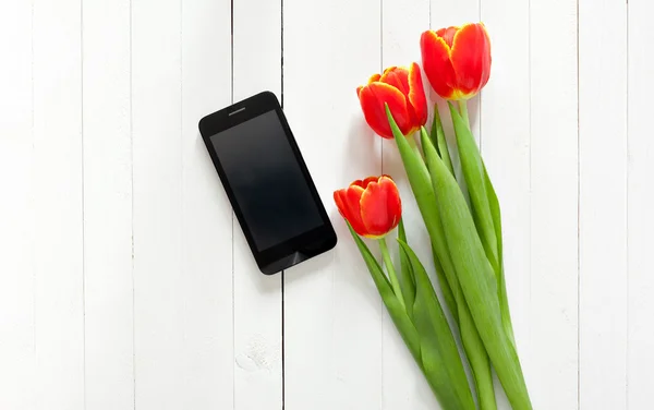 Spring bouquet of red tulips and black mobile phone on a white wooden background with copy space. View from the top with space for signature.
