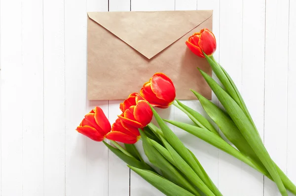 View from the top with space for signature. Spring bouquet of red tulips and a card in an envelope on a white wooden background with copy space.