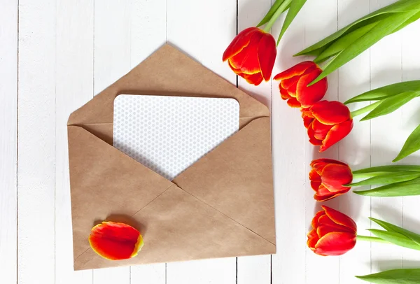 Spring bouquet of red tulips and a card in an envelope on a white wooden background with copy space. View from the top with space for signature.