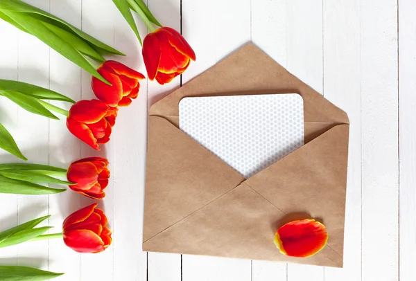 Spring bouquet of red tulips and a card in an envelope on a white wooden background with copy space. View from the top with space for signature.