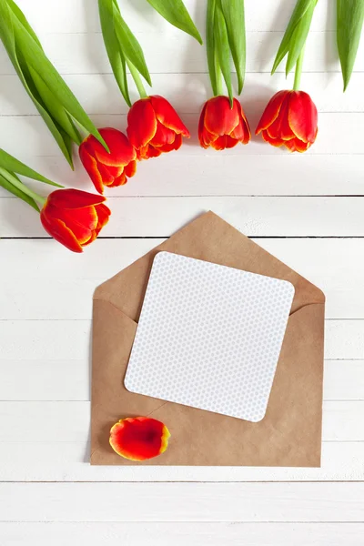 Spring bouquet of red tulips and a card in an envelope on a white wooden background with copy space. View from the top with space for signature.