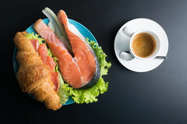 Croissant with salmon, lettuce on blue plate and cup of coffee o