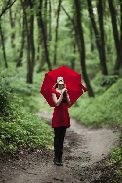Happy girl in red dress walking in fresh green forest with umbrella. Rain concept.