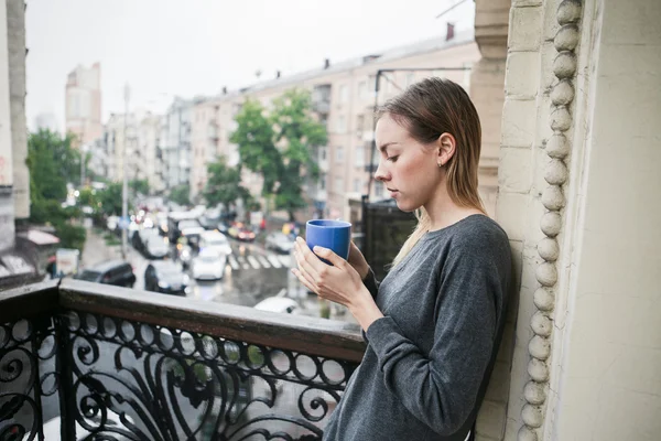 Beautiful young girl with a cup of morning coffee standing on the balcony in the rain. Thinking concept.
