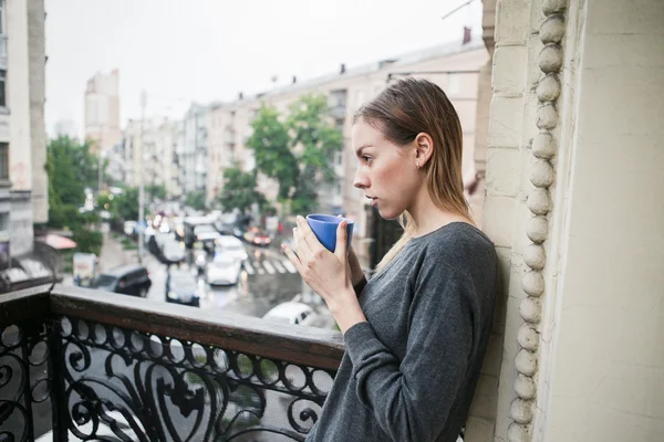 Beautiful young girl with a cup of morning coffee standing on the balcony in the rain. Thinking concept.