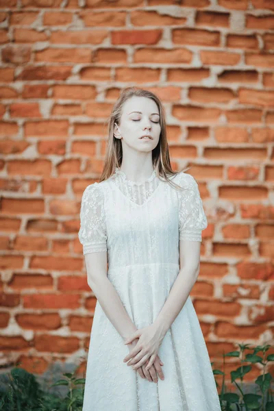 Beautiful young lady in white vintage dress standing near old brick wall