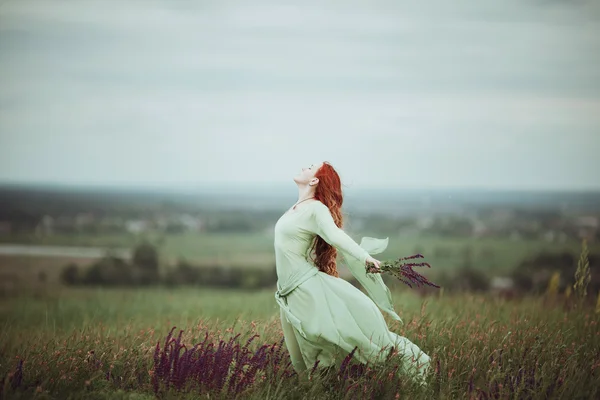 Young redhead girl in medieval dress walking through field with sage flowers. Wind concept. Fantasy