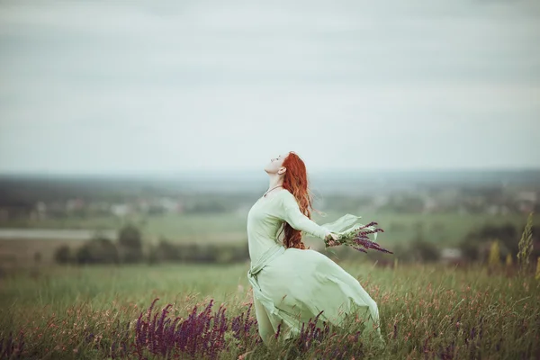 Young redhead girl in medieval dress walking through field with sage flowers. Wind concept. Fantasy