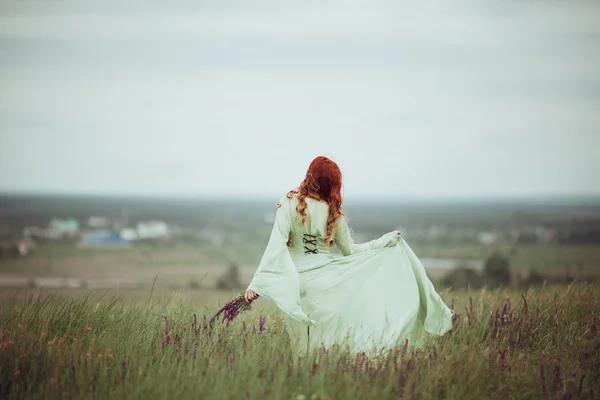 Young redhead girl in medieval dress walking through field with sage flowers. Wind concept. Fantasy